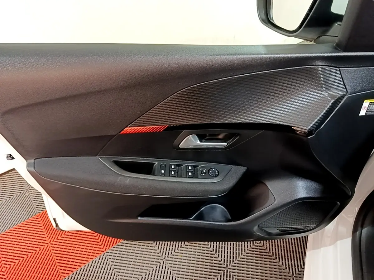 Vue intérieure du panneau de porte avant gauche de la Peugeot 208 électrique blanche, avec commandes de vitres et insert effet carbone.