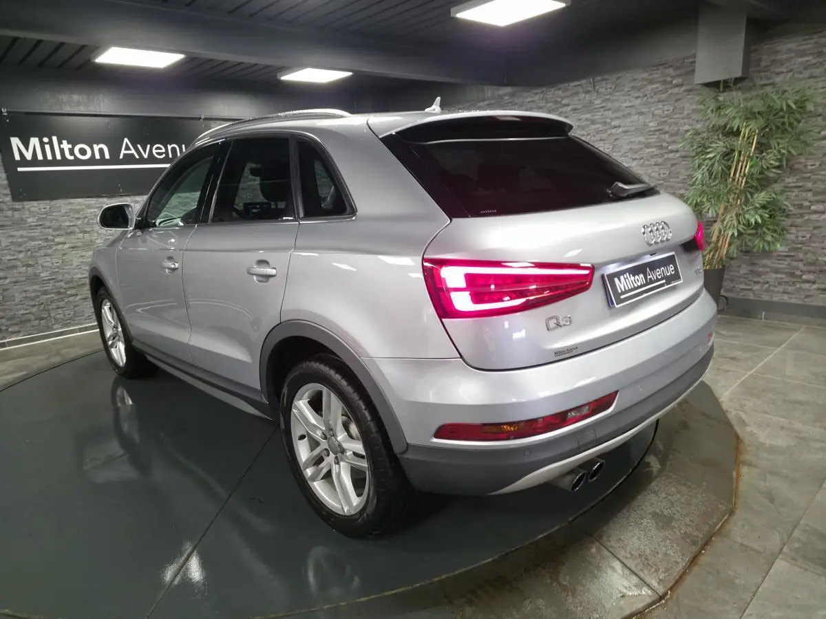 Audi Q3 gris métallisé vue 3/4 arrière droit avec feux arrière allumés dans un showroom intérieur.