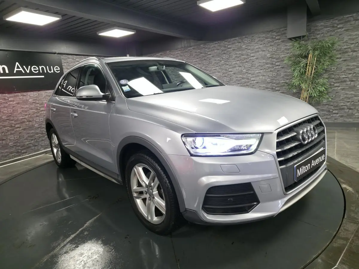 Audi Q3 gris métallisé en 3/4 avant droit, avec calandre Singleframe et phares LED allumés dans un showroom.