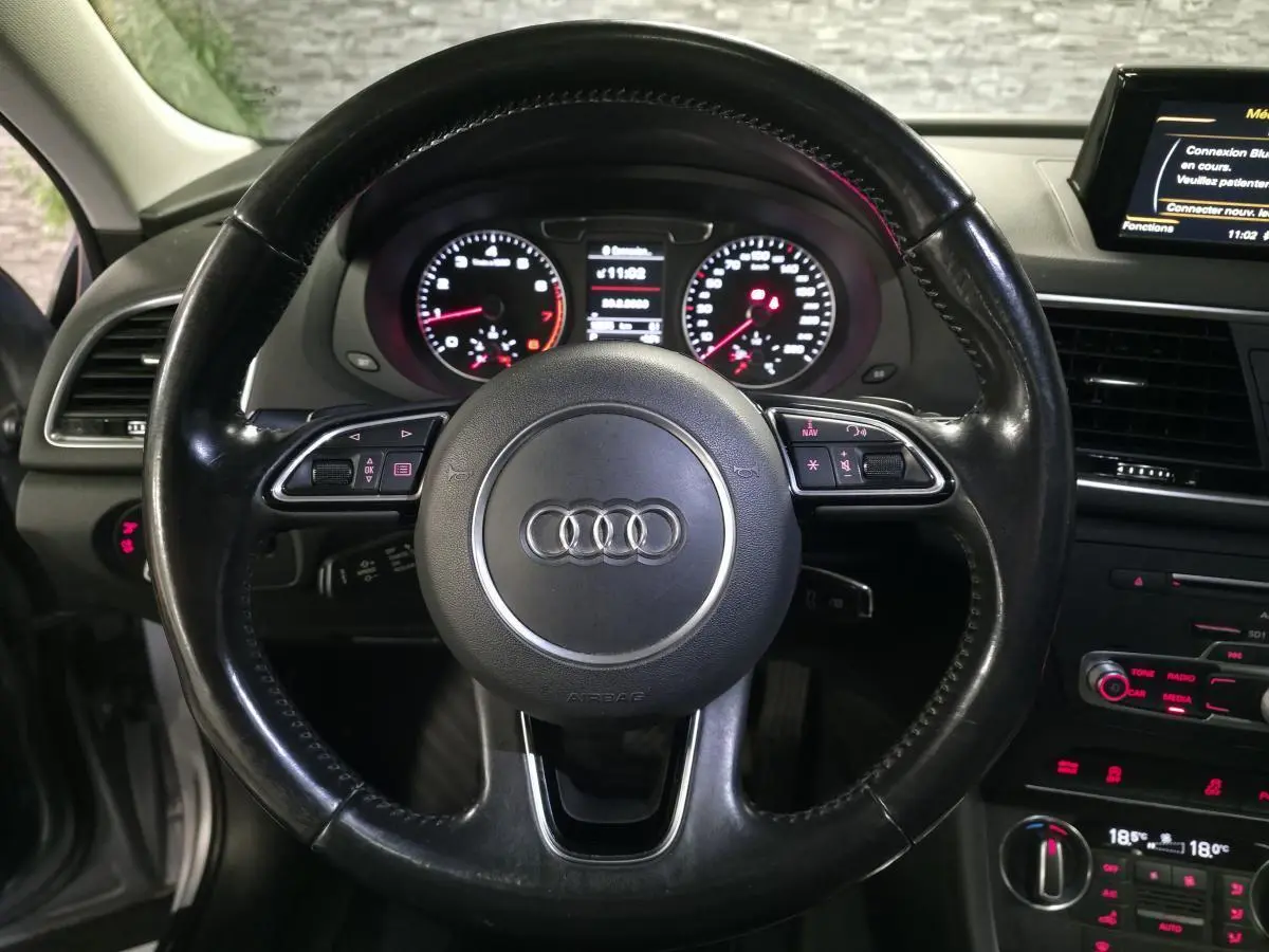 Vue intérieure centrée sur le volant en cuir noir avec logo Audi, tableau de bord et console centrale du Q3 gris de 2016.