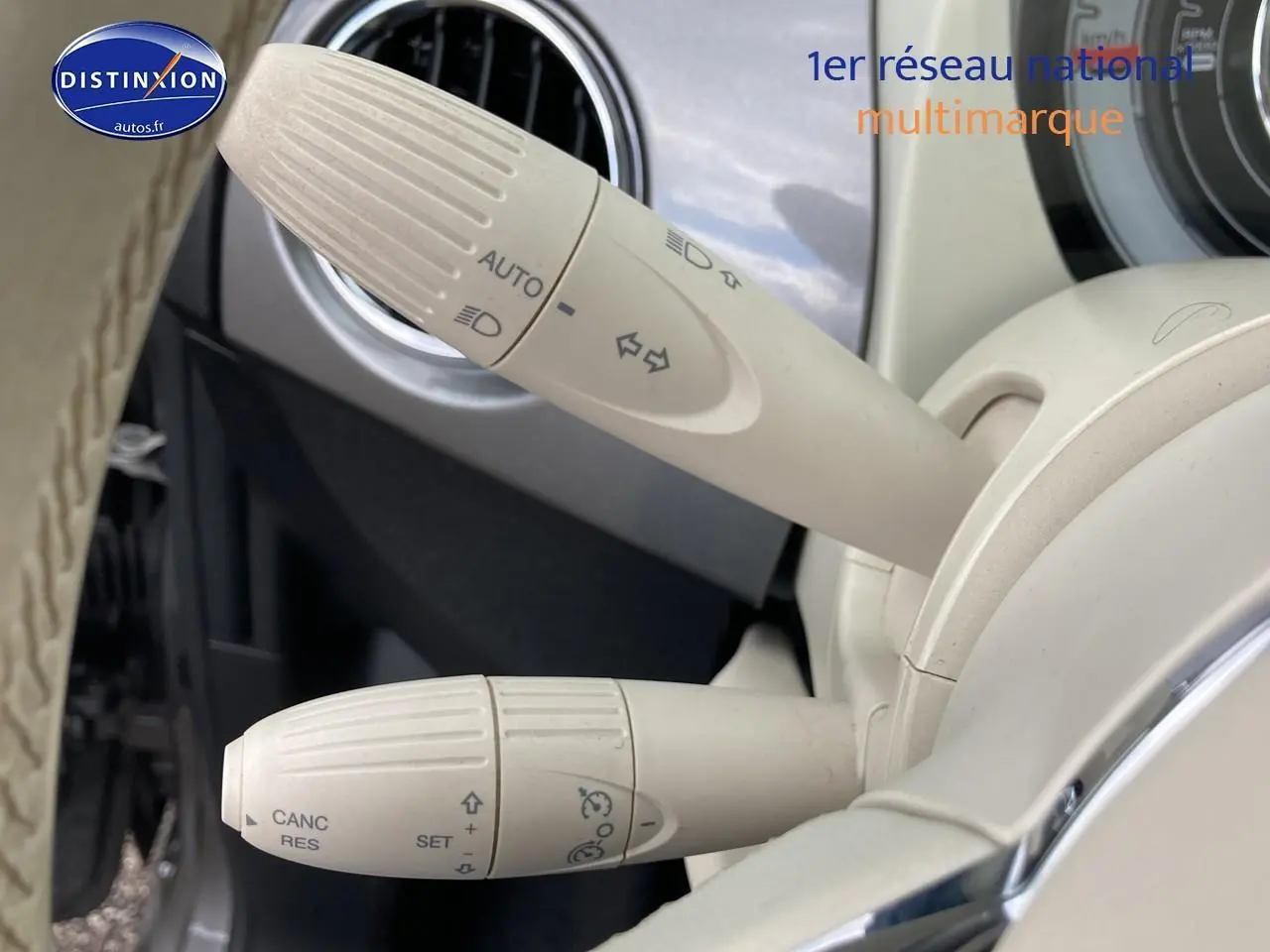 Gros plan sur les commandes blanches du volant d’une FIAT 500 gris, mettant en valeur les fonctions de régulateur et feux automatiques.
