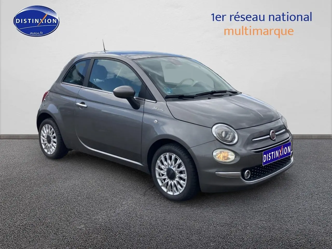 Fiat 500 gris vue 3/4 avant droit, avec jantes alu et phares allumés sur fond neutre.