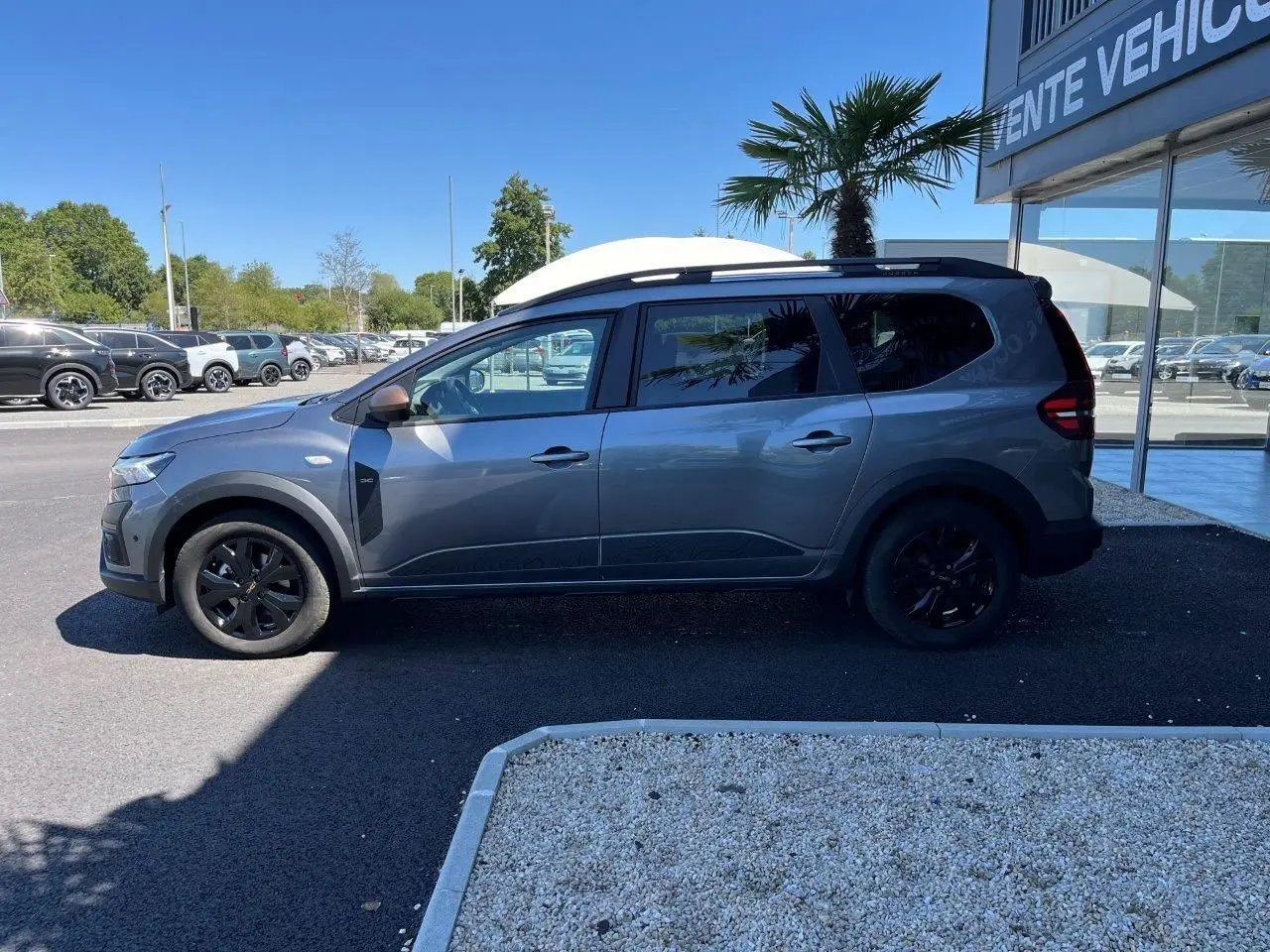 Profil droit du Dacia Jogger 1.6 Hybrid gris schiste 2025 avec jantes noires et toit noir, stationné devant un showroom.