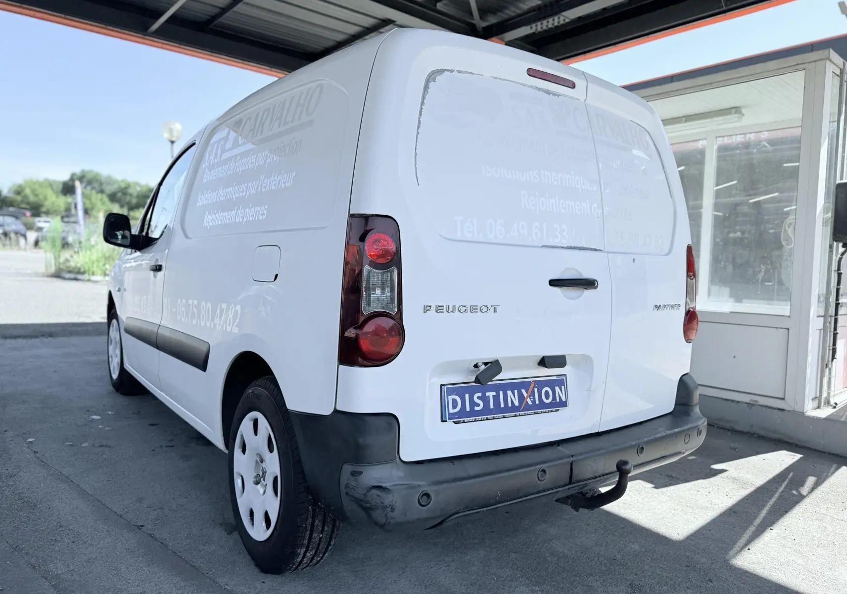 Vue 3/4 arrière droite d’un Peugeot Partner blanc utilitaire avec marquage publicitaire et attelage de remorque.