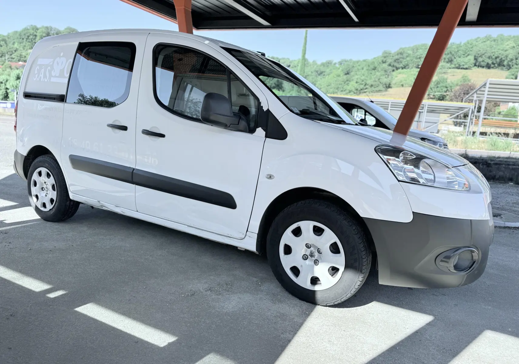 Peugeot Partner blanc en 3/4 avant droit, avec protections noires sur les portières et jantes acier sous abri.
