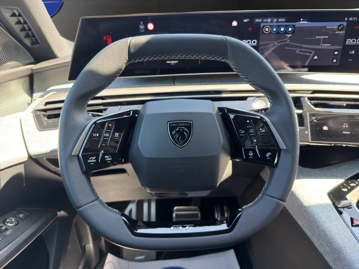 Vue rapprochée du volant cuir perforé noir avec logo Peugeot et inscription GT, tableau de bord numérique en arrière-plan.