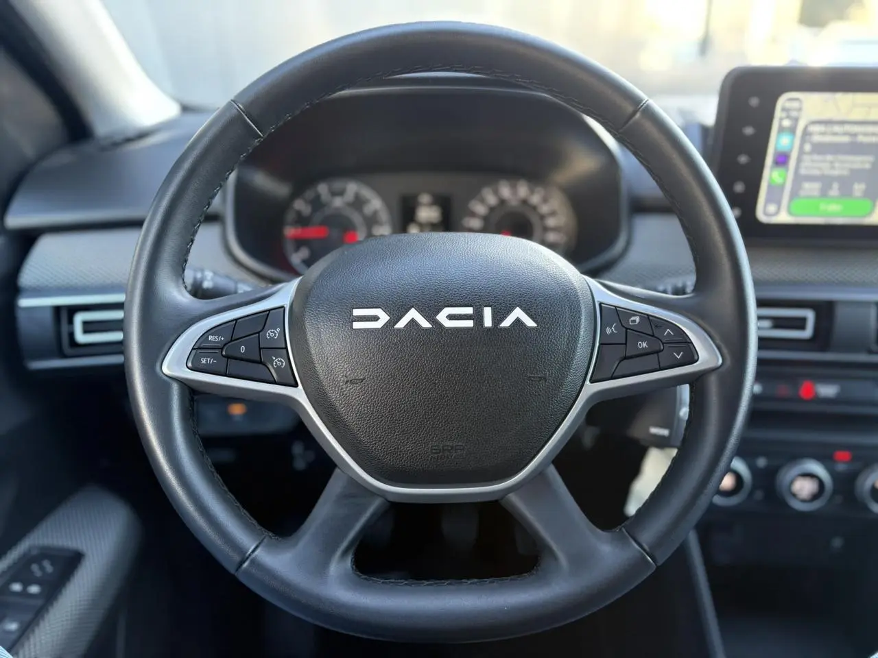 Gros plan sur le volant noir de la Dacia Jogger 2025 avec commandes intégrées et tableau de bord flou en arrière-plan.