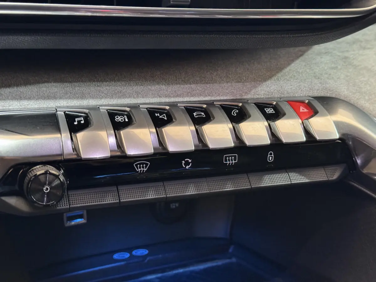 Gros plan sur la rangée de boutons chromés et noirs du tableau de bord du Peugeot 3008 gris clair, avec commandes clim et hazard.