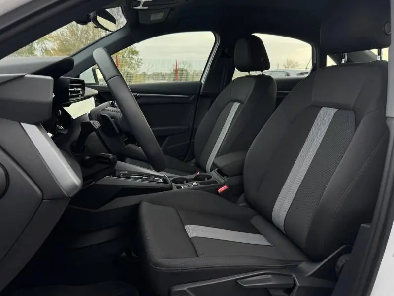 Vue intérieure côté gauche de l'Audi A3 Sportback 2024, sièges tissu noir avec bande grise centrale et volant multifonction.