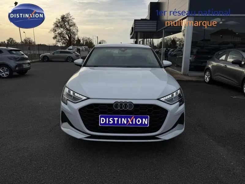 Vue frontale d'une Audi A3 Sportback blanche 2024 avec calandre noire et phares LED allumés sur parking.