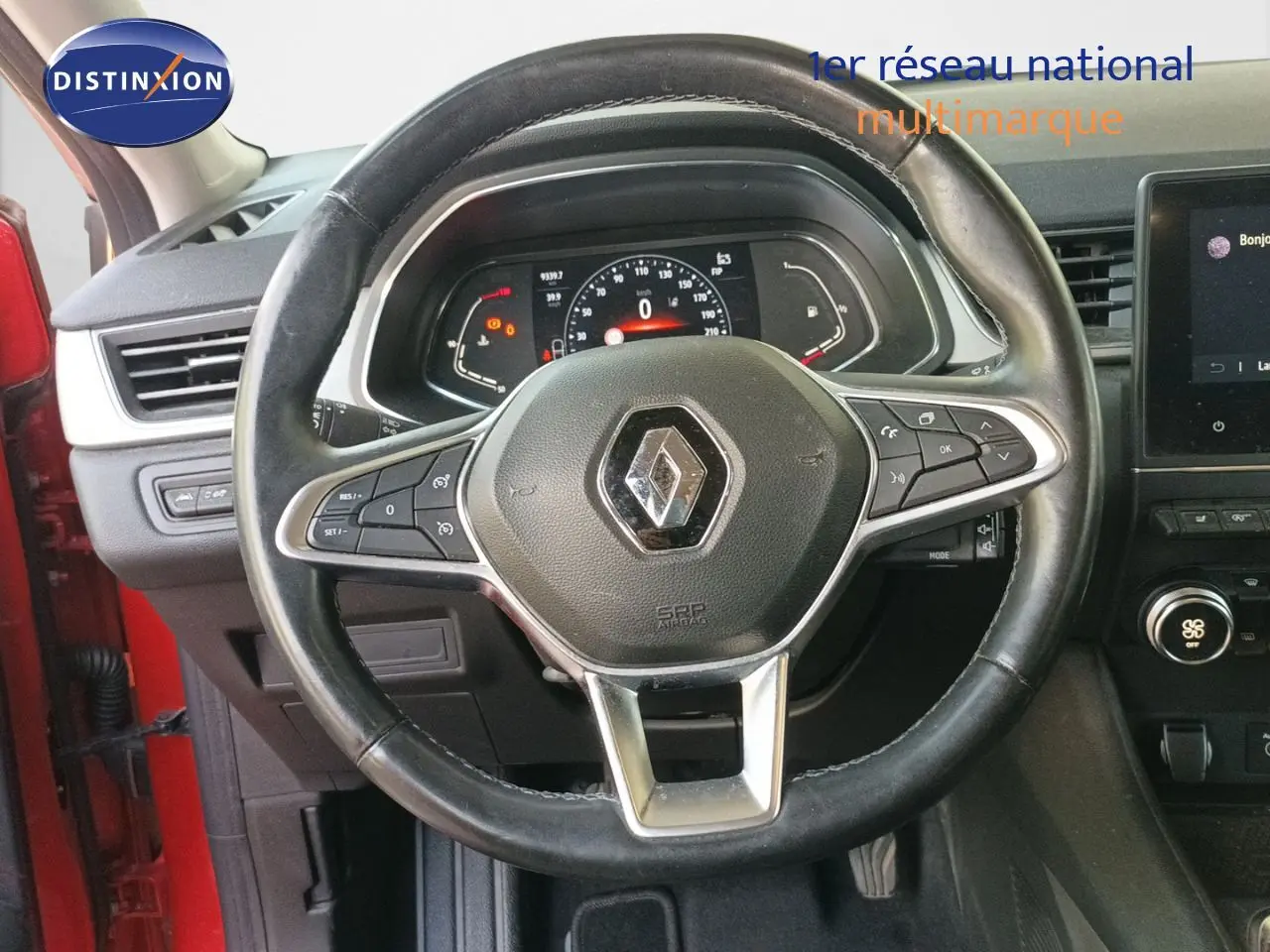 Vue intérieure centrée sur le volant noir avec logo Renault, tableau de bord digital et partie rouge de la portière visible.