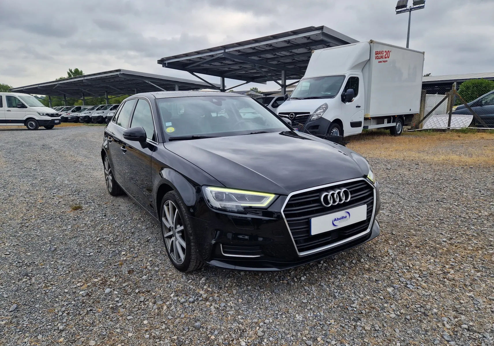 Audi A3 Sportback noire vue 3/4 avant droit sur un sol gravillonné avec des véhicules utilitaires en arrière-plan.