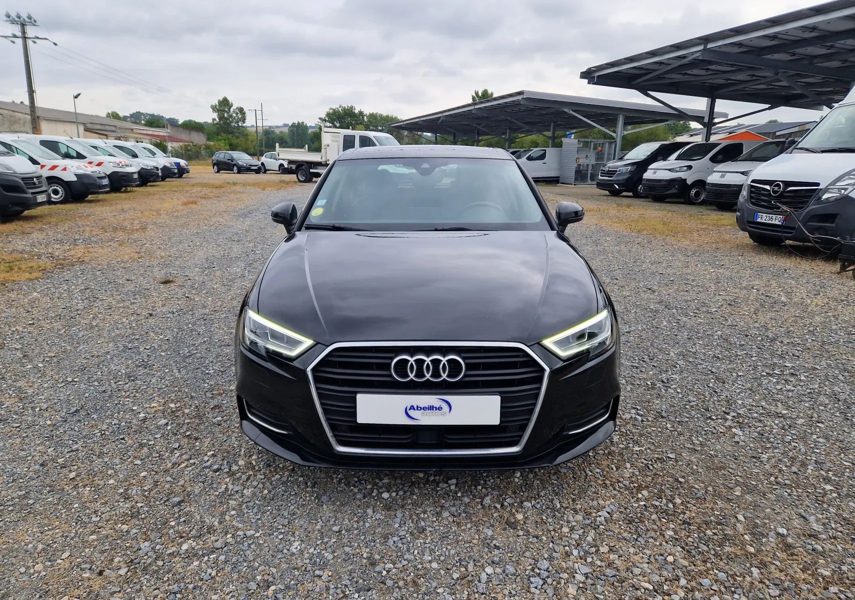 Vue de face d'une Audi A3 Sportback noire de 2019 avec phares allumés sur un parking gravillonné.