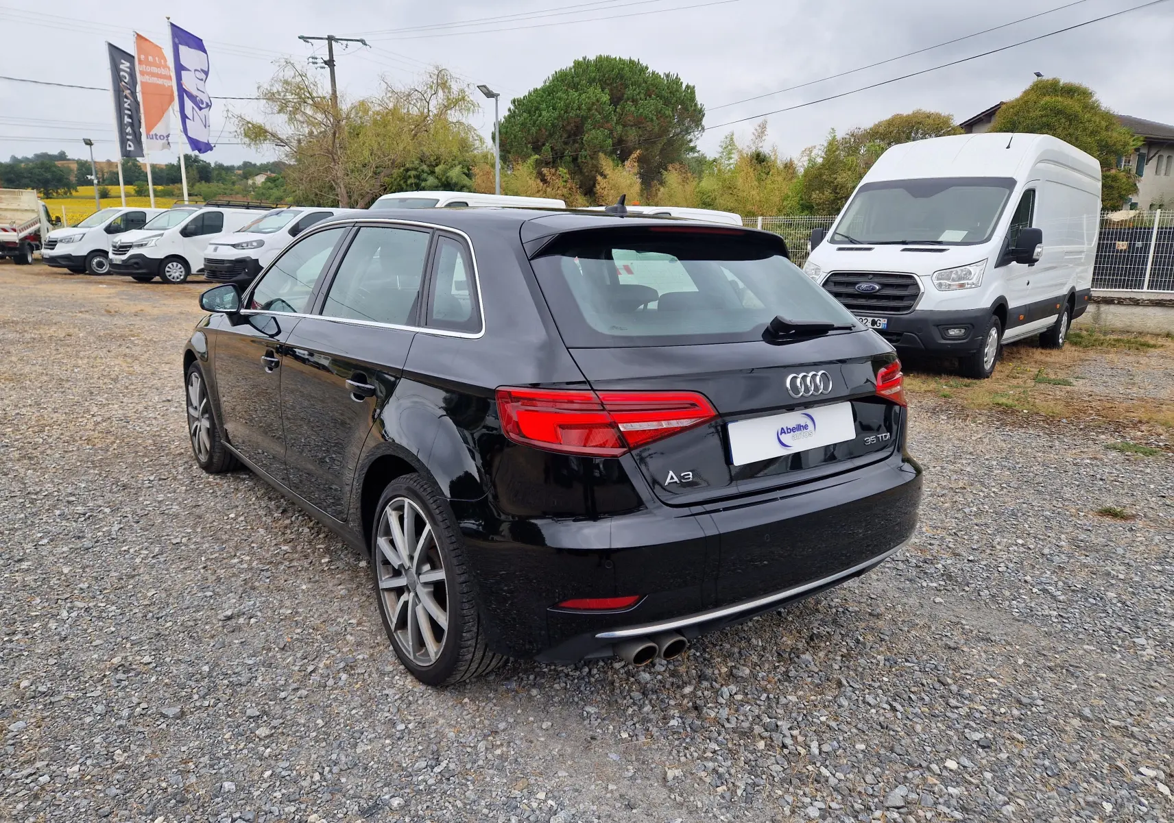 Audi A3 Sportback noir vue 3/4 arrière droit sur parking avec plusieurs véhicules utilitaires blancs en arrière-plan.