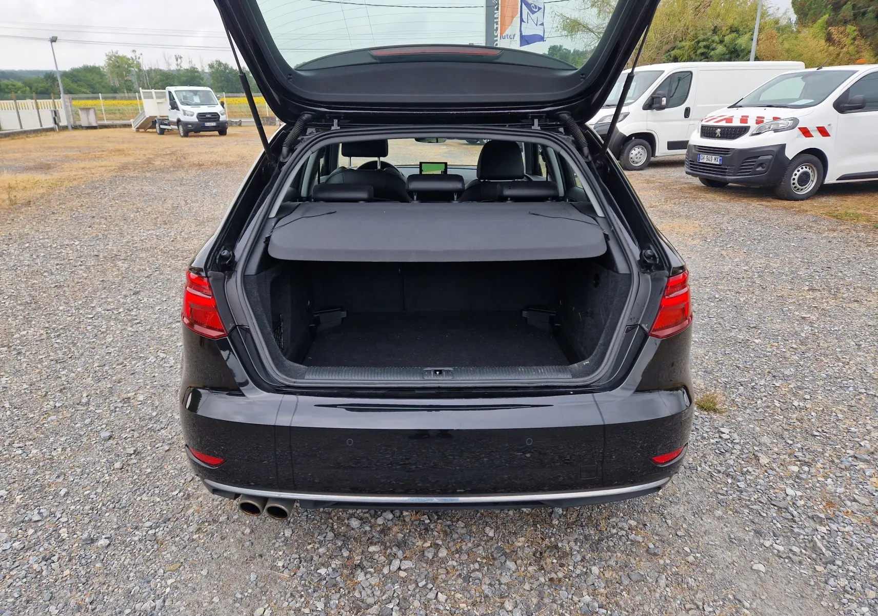 Vue arrière d'Audi A3 Sportback noire 2019 avec coffre ouvert et cache-bagages en place sur un sol gravillonné.