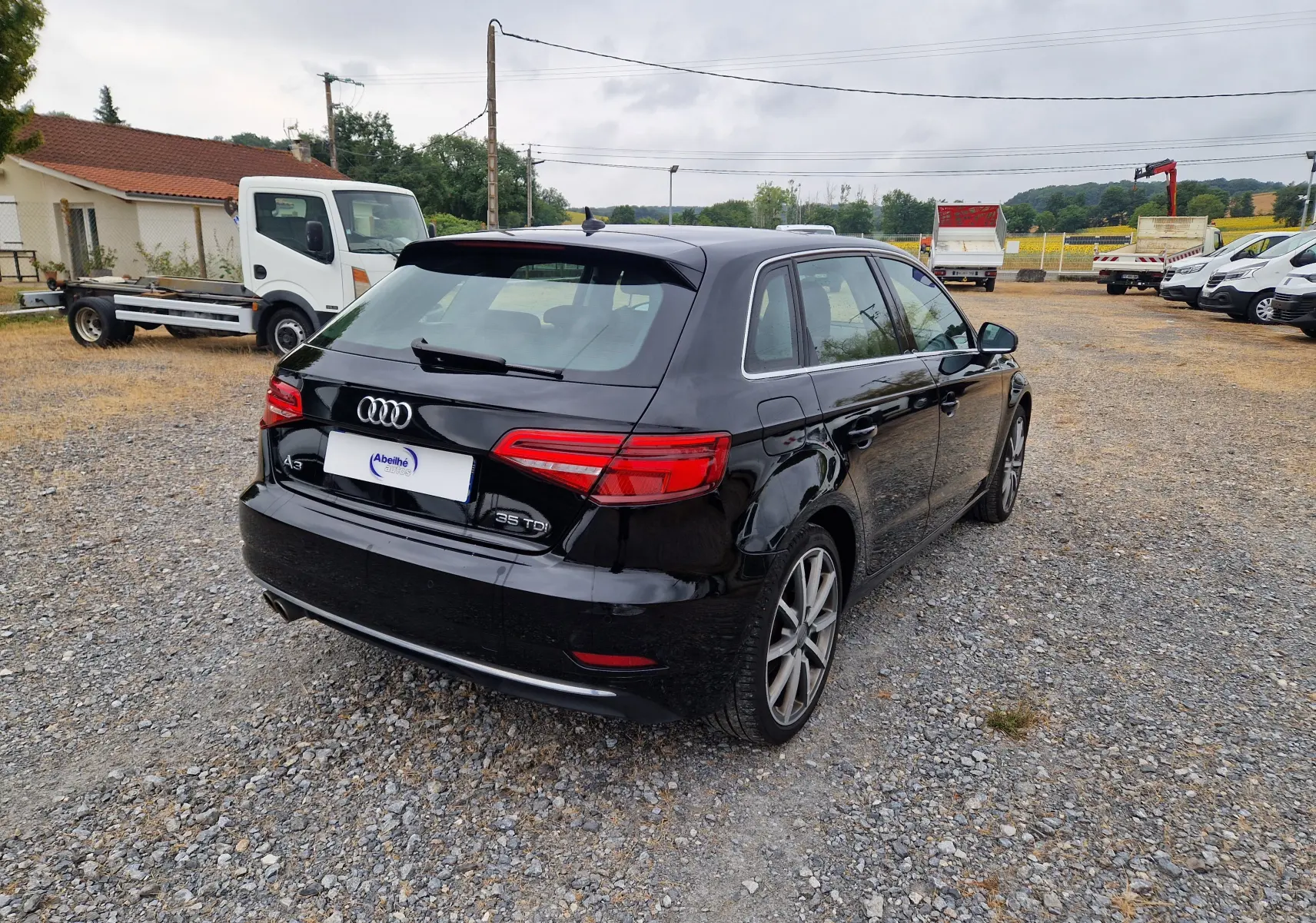 Audi A3 Sportback noir vue 3/4 arrière droit sur un sol gravillonné avec plusieurs véhicules en arrière-plan.