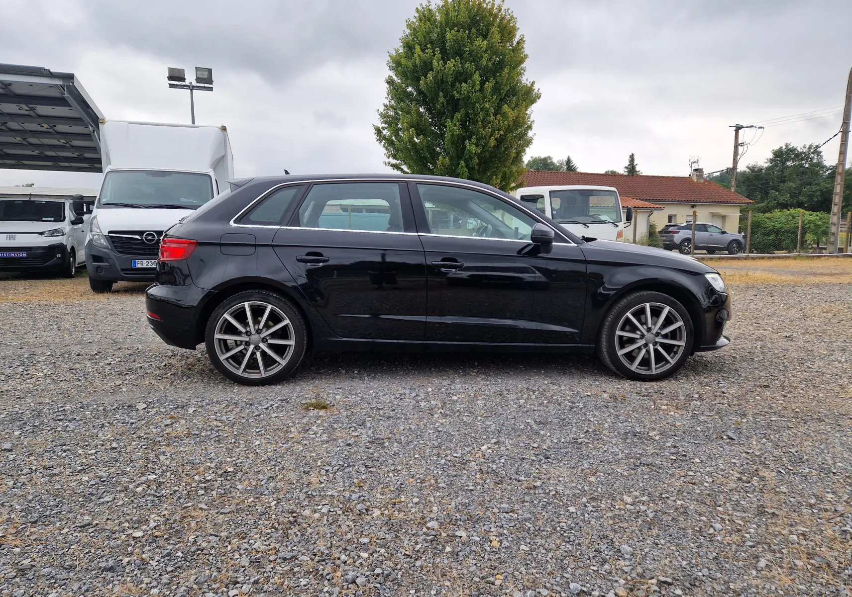 Audi A3 Sportback noir vue de profil côté gauche, sur un sol gravillonné avec des véhicules en arrière-plan.