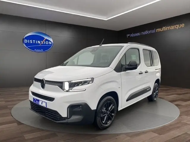 Vue 3/4 avant droite d'un Citroën Berlingo blanc 2025 avec phares allumés et jantes noires en showroom.