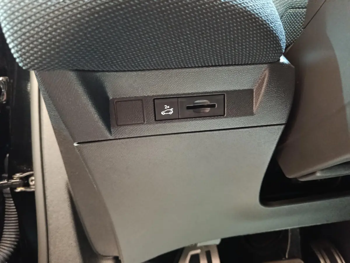 Détail intérieur montrant le bouton d'ouverture électrique du coffre sous le siège avant, plastique noir texturé.