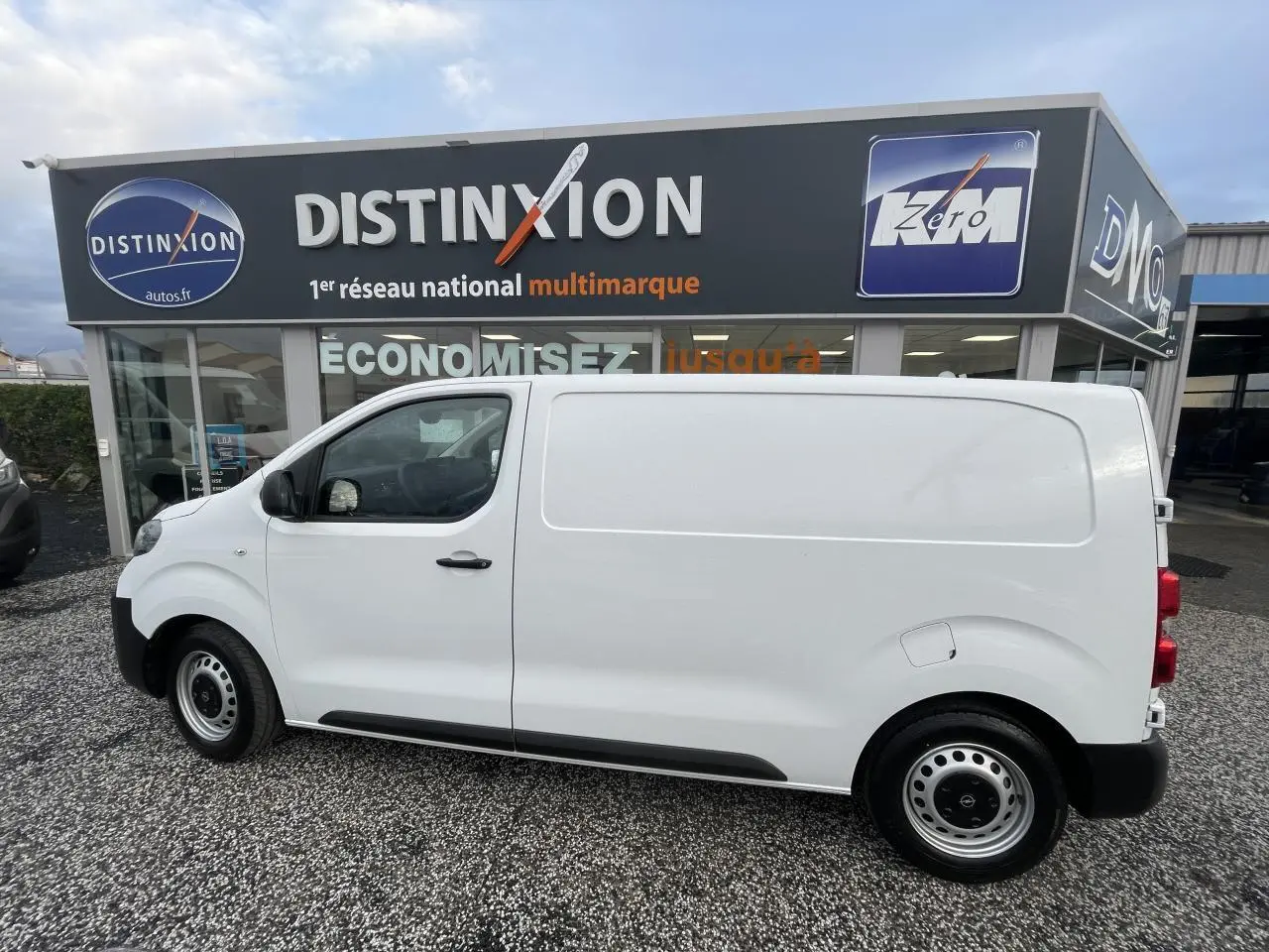 Vue de profil côté gauche d'un utilitaire Opel Vivaro blanc stationné devant un concessionnaire multimarque.