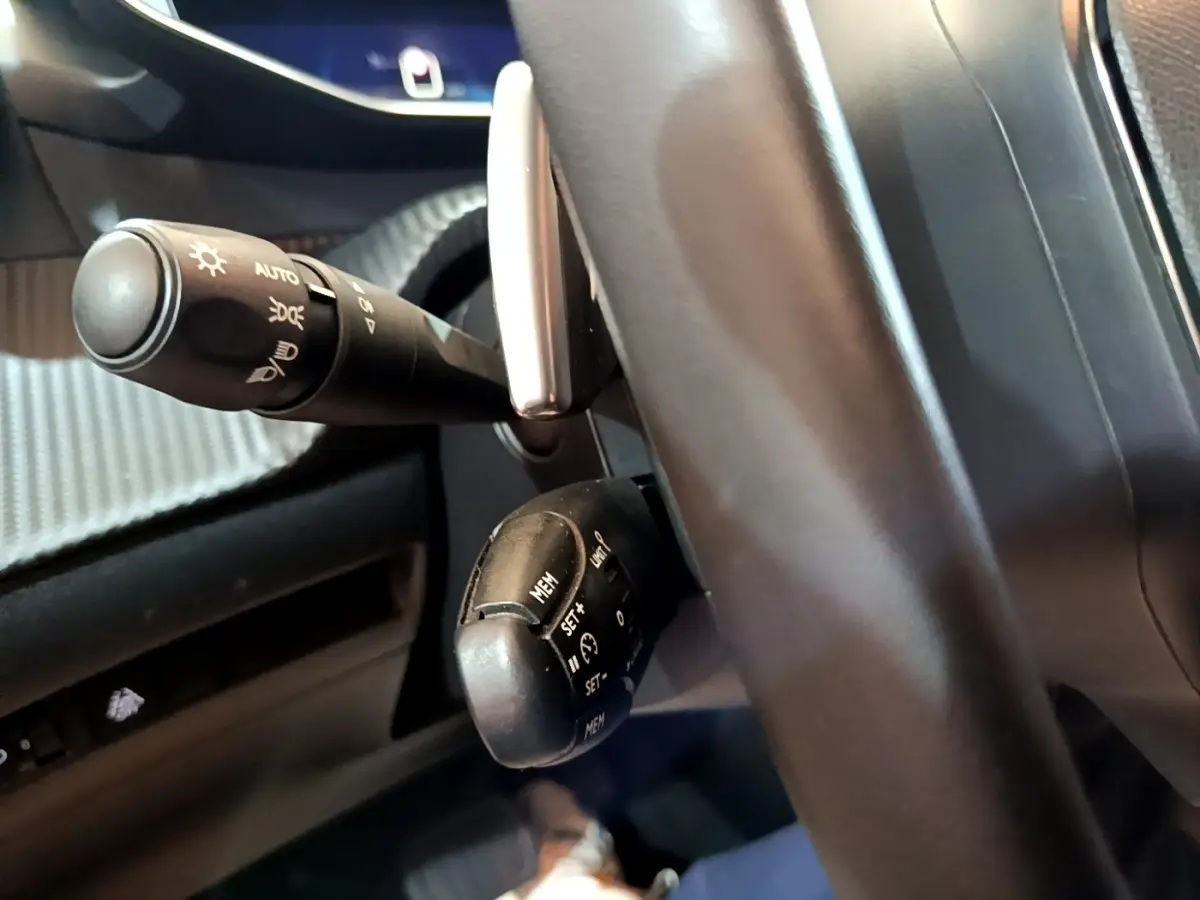 Gros plan sur la commande de régulateur de vitesse et le commodo d'éclairage à gauche du volant noir dans une Peugeot 208 jaune.