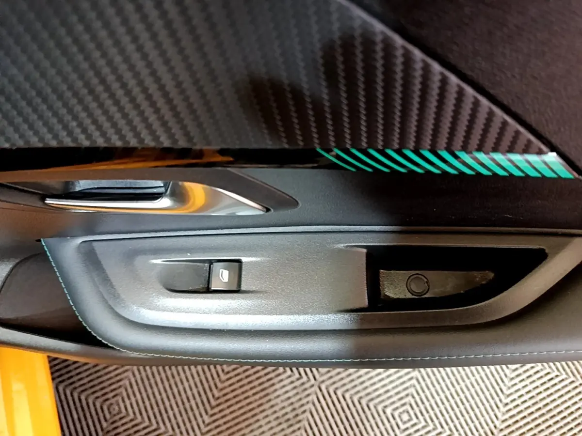 Gros plan sur la poignée intérieure côté gauche d’une Peugeot 208 jaune avec surpiqûres bleues et insert carbone.