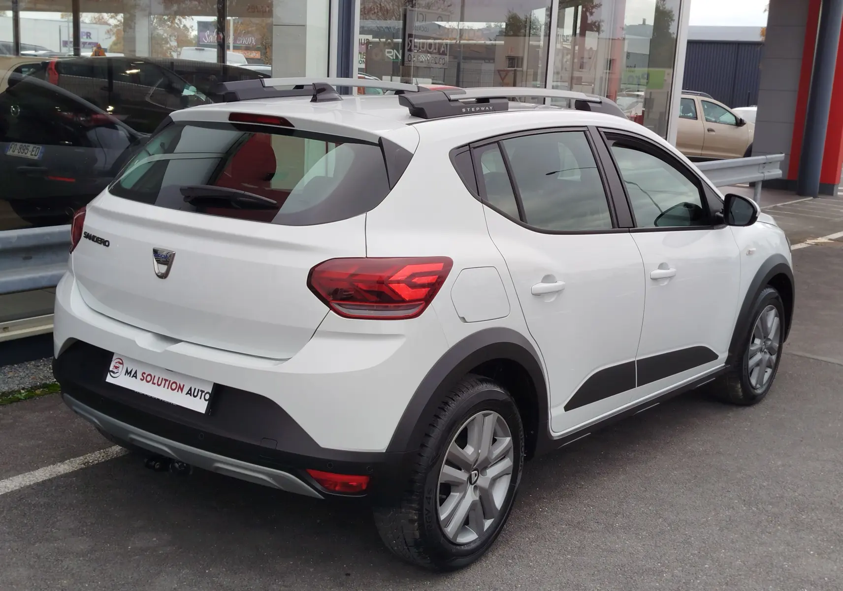 Vue 3/4 arrière droite d'une Dacia Sandero Stepway blanche glacier avec barres de toit et protections noires sur les bas de caisse.