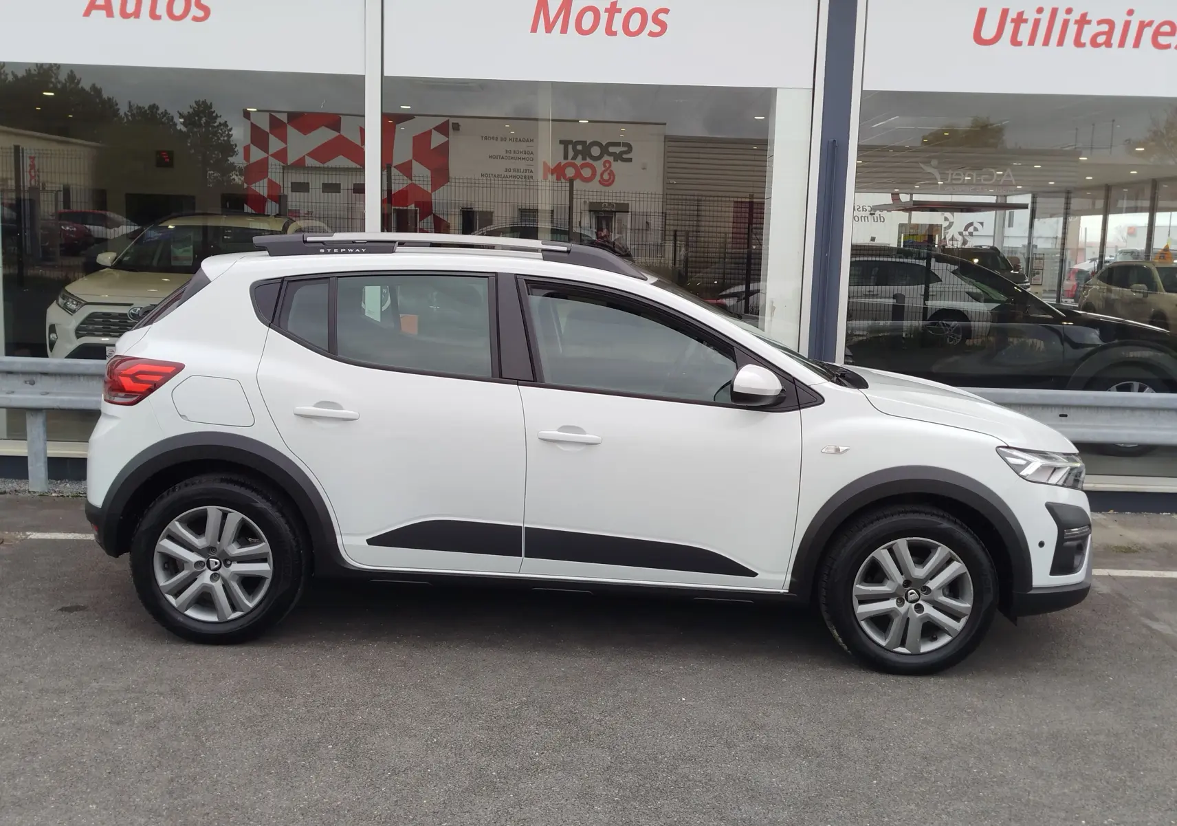 Vue latérale droite d'une Dacia Sandero Stepway blanche Glacier avec barres de toit et protections noires sur les bas de caisse.