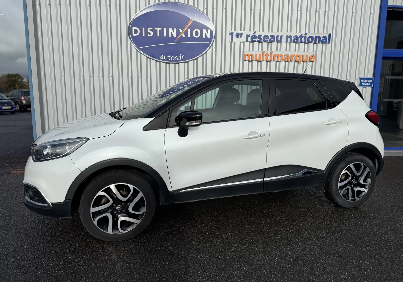 Profil côté gauche d'un Renault Captur blanc 2017 avec toit noir et jantes alliage bicolores devant un bâtiment Distinxion.