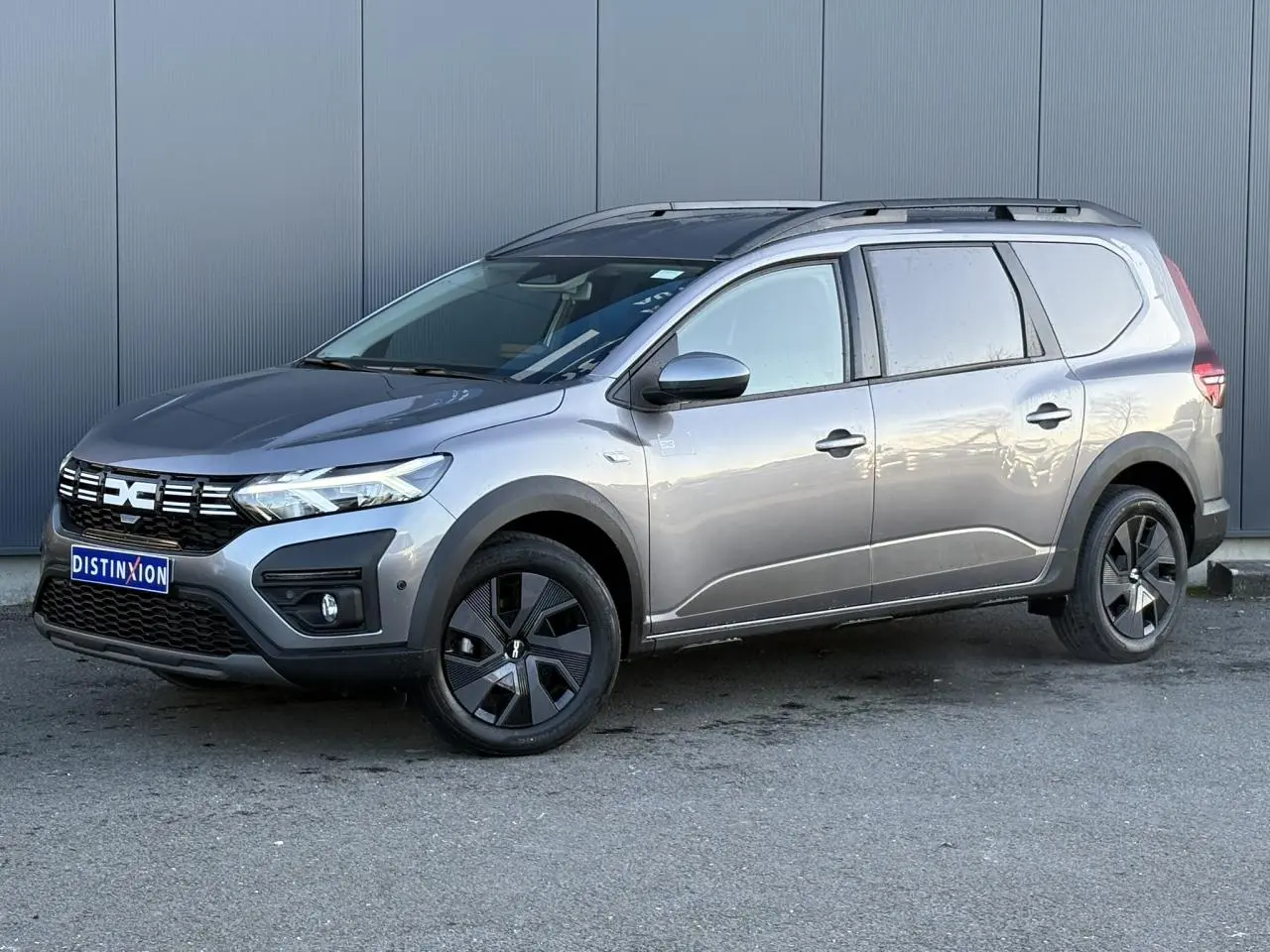 Vue 3/4 avant droite d'un Dacia Jogger gris schiste avec barres de toit et jantes noires distinctives.