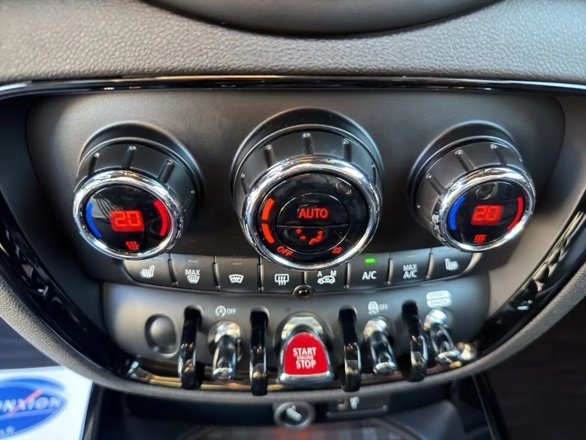 Gros plan sur la console centrale noire de la MINI Clubman blanche, avec commandes de climatisation et bouton rouge Start/Stop.