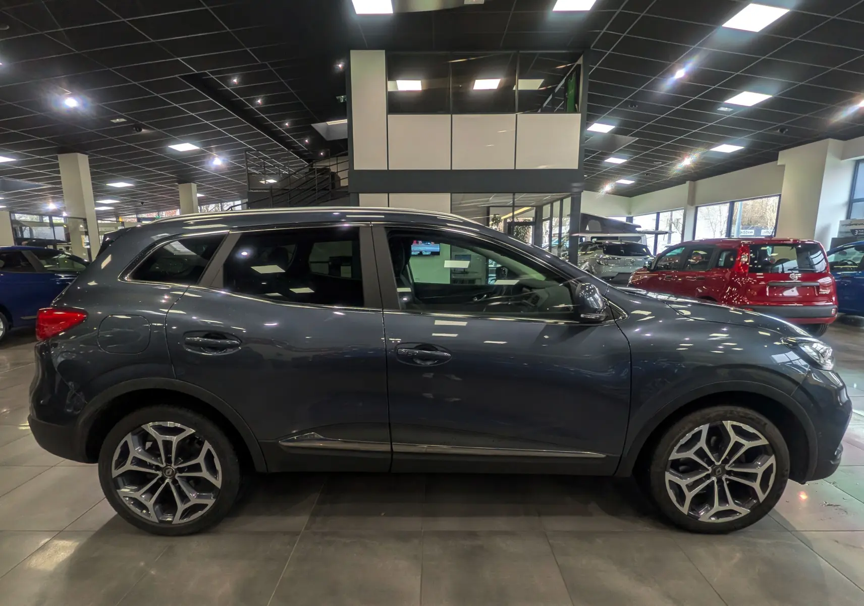 Profil côté gauche d’un Renault Kadjar gris titanium 2019 en showroom, avec jantes alliage et vitres teintées.
