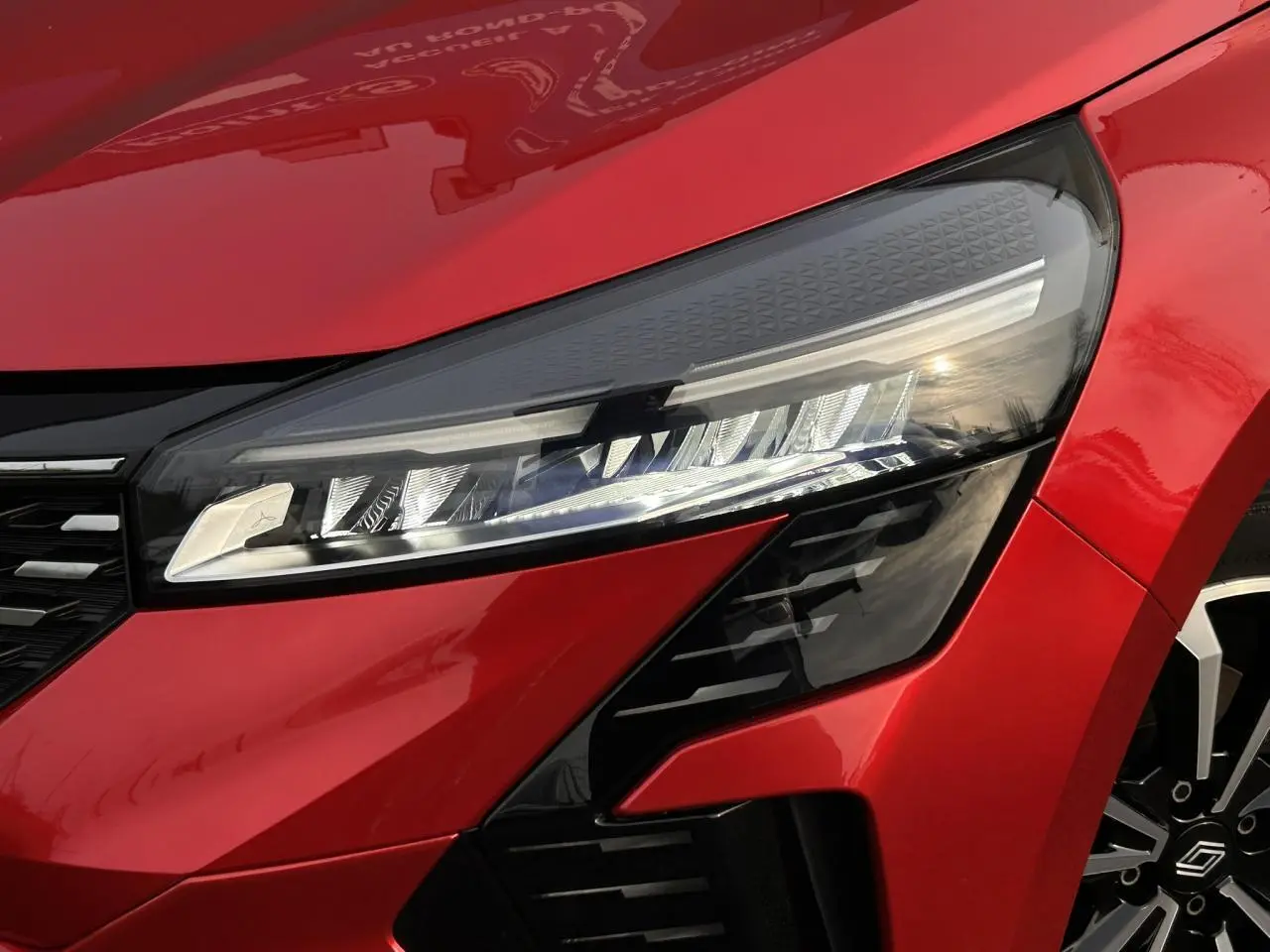 Gros plan sur le phare avant droit et la calandre d'une Renault Clio 2025 Rouge Désir avec jante alliage visible.
