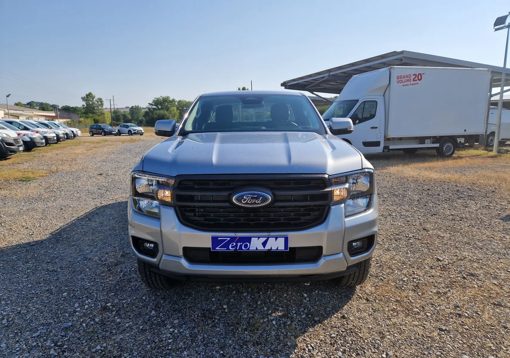 Vue avant frontale du Ford Ranger Super Cabine gris Iconic, avec calandre noire et phares allumés sur terrain gravillonné.