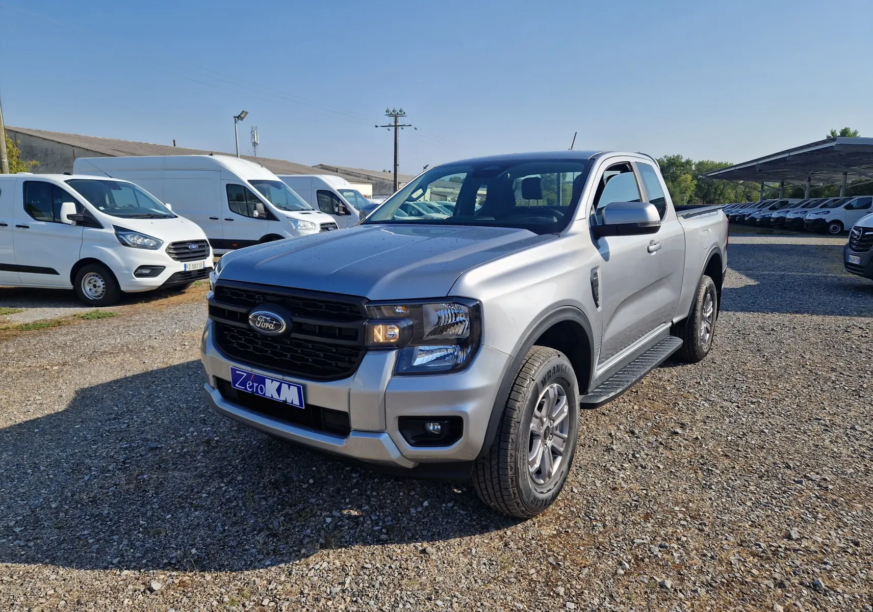 Ford Ranger Super Cabine gris iconic vu en 3/4 avant droit sur un terrain gravillonné avec plusieurs véhicules utilitaires blancs en arrière-plan.