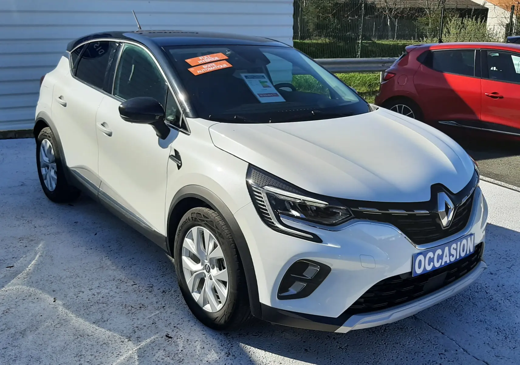 Vue 3/4 avant droite d’un Renault Captur blanc nacré avec toit noir, affichant une plaque "OCCASION" sur un parking extérieur.