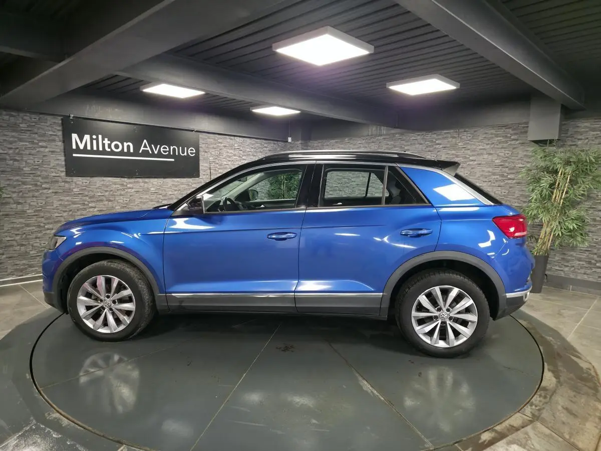 Vue de profil côté gauche d'un Volkswagen T-Roc bleu foncé avec jantes alliage et toit noir en intérieur showroom.