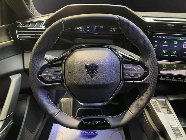 Vue rapprochée du volant cuir noir de la Peugeot 308 Hybrid 145 e-DCS6 GT avec commandes multifonctions et logo central.