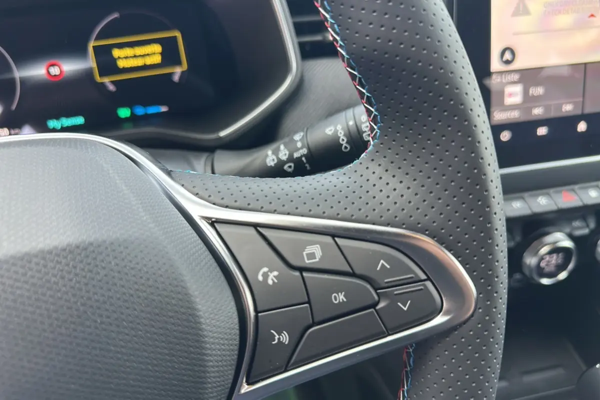 Gros plan sur les commandes multifonctions du volant cuir perforé de la Renault Clio V E-Tech gris rafale, tableau de bord en arrière-plan.