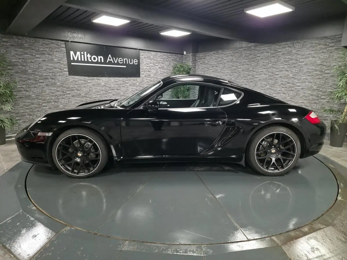 Profil droit d'une Porsche Cayman 2.7i noire de 2008 avec jantes SportDesign noires de 19 pouces, en intérieur showroom.