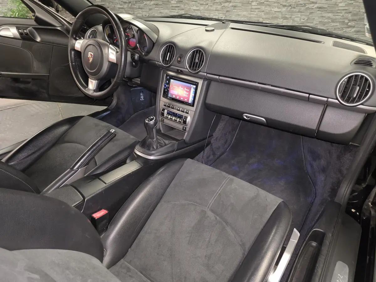 Vue intérieure côté passager d’une Porsche Cayman 2.7i noire de 2008 avec tableau de bord noir et levier de vitesses manuel.