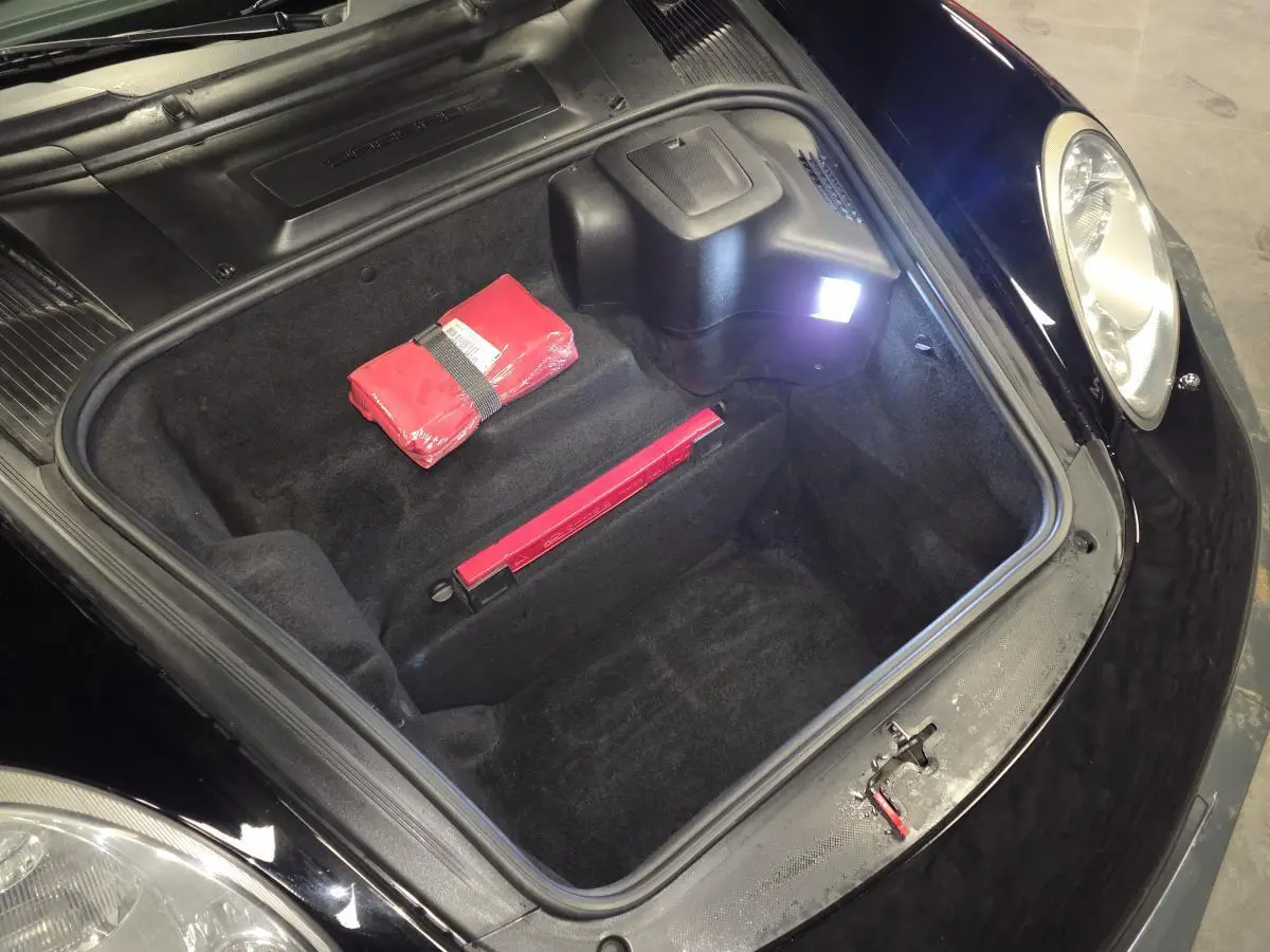 Vue plongeante sur le coffre avant noir d'une Porsche Cayman 2.7i 2008 avec trousse de secours rouge visible.