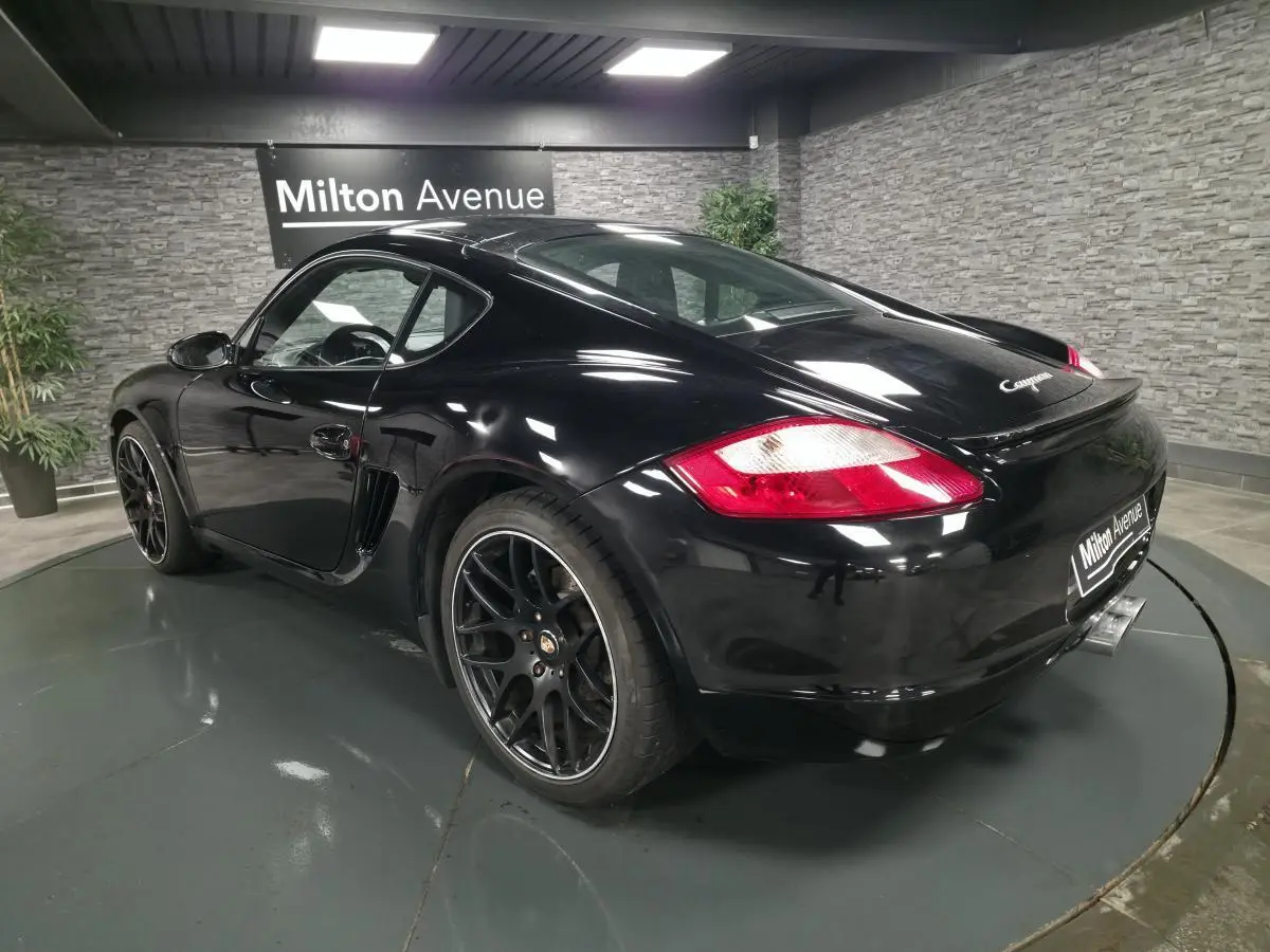Vue 3/4 arrière droite d'une Porsche Cayman 2.7i noire avec jantes SportDesign noires de 19 pouces et double sortie d'échappement centrale.