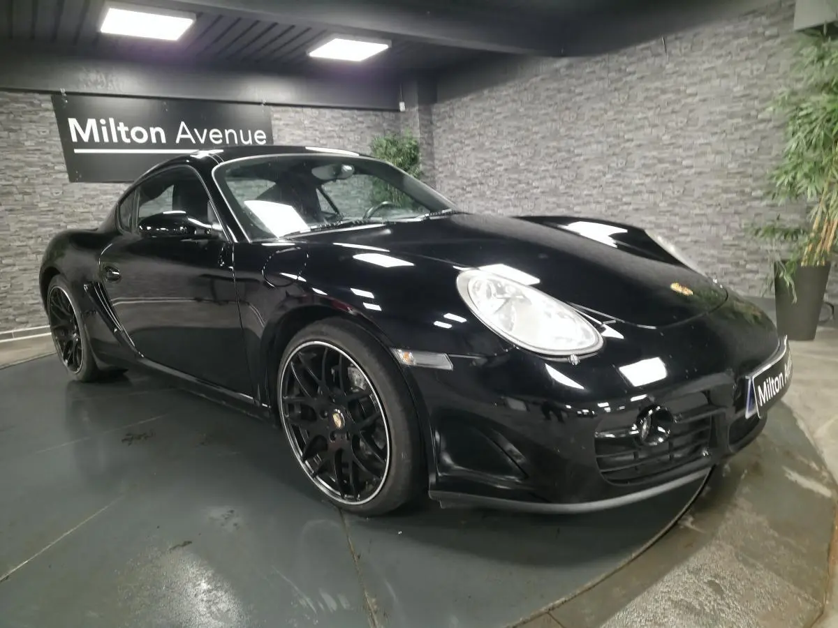 Porsche Cayman 2.7i noire vue 3/4 avant droit avec jantes SportDesign noires de 19 pouces dans un showroom.