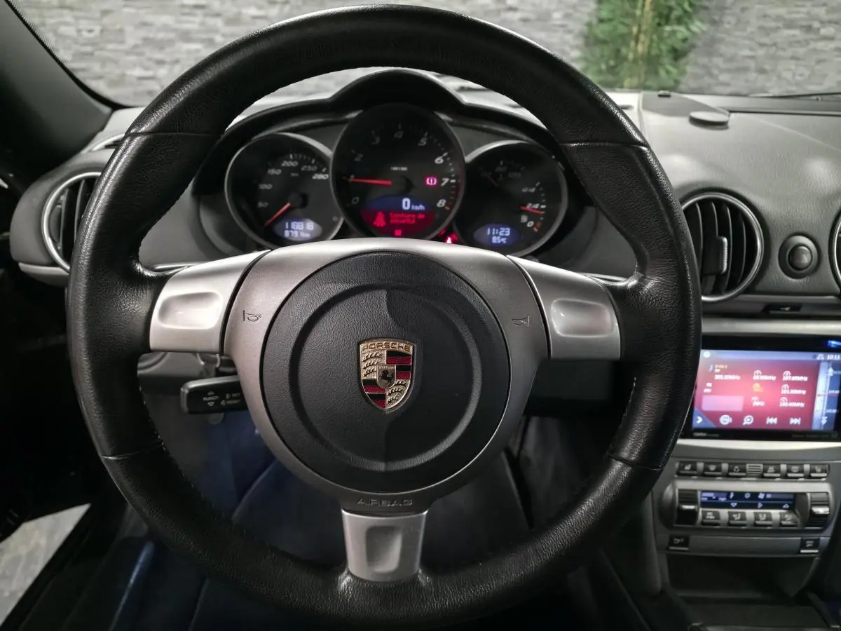 Vue intérieure centrée sur le volant noir en cuir et le tableau de bord avec compteurs ronds du Porsche Cayman 2.7i 2008.