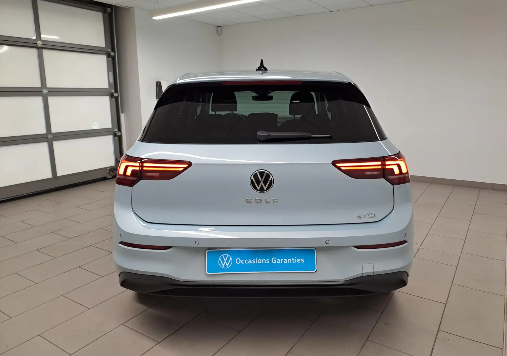 Vue arrière d'une Volkswagen Golf 1.5 eTSI 116 DSG7 Bleu Glacier avec feux allumés dans un garage.