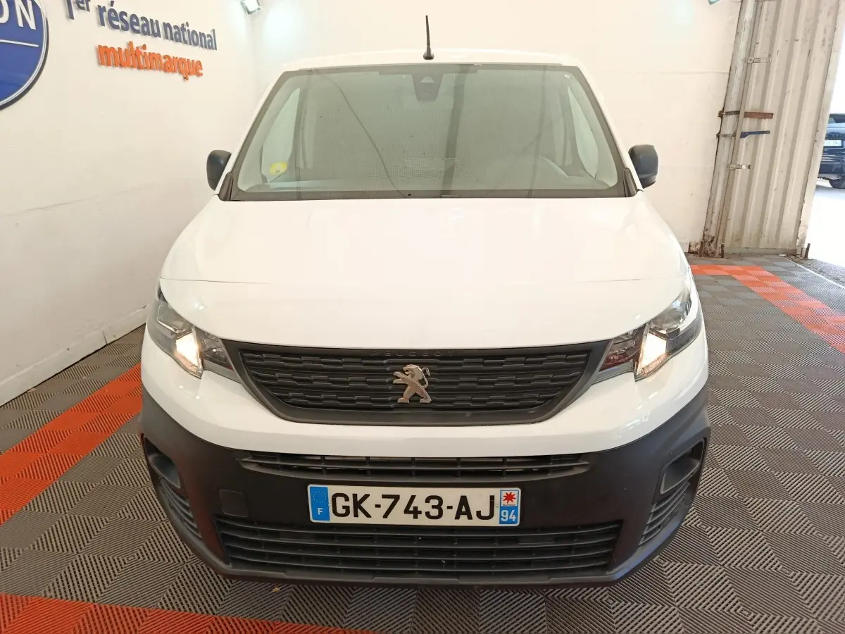 Vue frontale d’un Peugeot Partner blanc 2022 avec calandre noire et phares allumés en intérieur showroom.