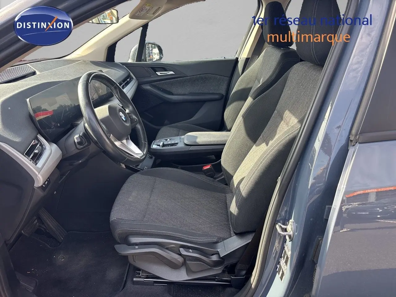 Vue intérieure côté conducteur d'une BMW Série 2 Active Tourer 218d 2023 avec sièges tissu gris et volant cuir, porte ouverte.