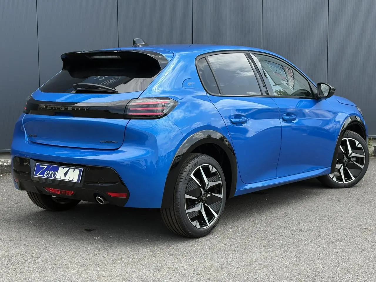 Vue 3/4 arrière droite d’une Peugeot 208 Hybrid 110 GT bleu Vertigo avec jantes alliage bi-tons et toit noir.