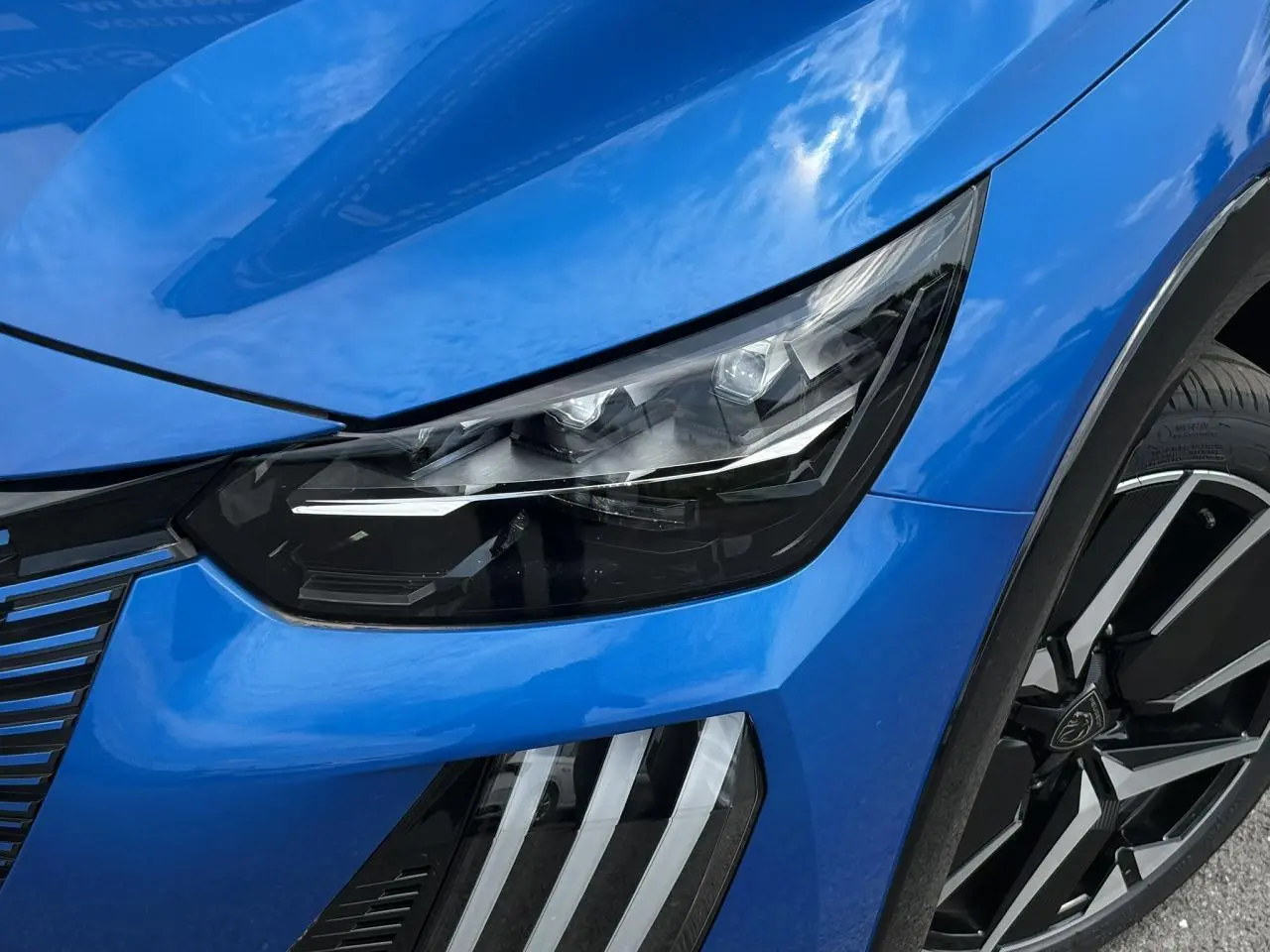 Gros plan sur l'avant droit bleu Vertigo du Peugeot 208 Hybrid 110 e-DSC6 GT, mettant en valeur le phare LED et la jante alliage diamantée.